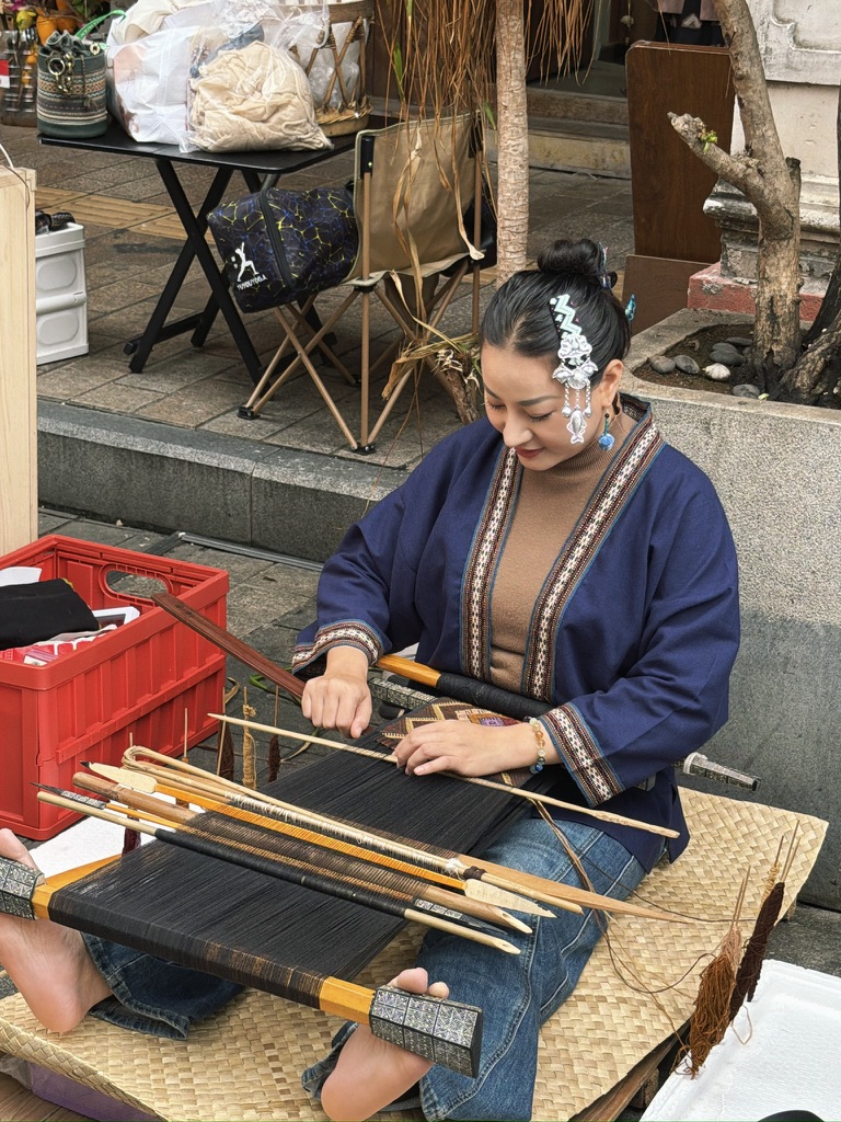 一位黎族的小姐姐在街边展示民族织布工艺,不过看她手脚并用地忙着织布,身边的摊子也照顾不到了。